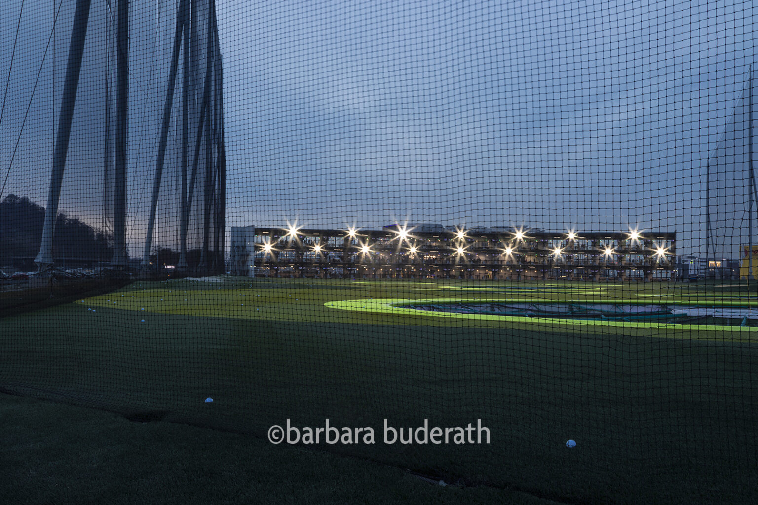 Fotografie: Sportanlage Topgolf in Oberhausen in der Abendstunde. Man sieht auf das Abschlagsgebäude. Fotografiert durch das Sicherungsnetz