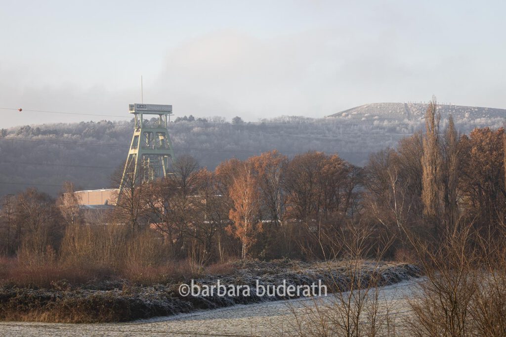 Fotografie: Blick auf den Förderturm der ehemaligen Zeche Prosper Haniel in Bottrop an einem kalten Wintertag. Hinter dem Förderturm sieht man eine mit Rasureif überzogene Halde.