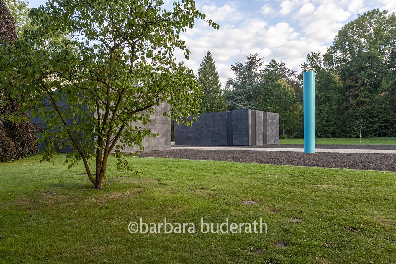 Aussenaufnahme der Rückseite des Museums unter Tage im Park von Haus Weitmar mit einem Strauch im linken Vordergrund und einer blauen Steele auf der rechten Seite