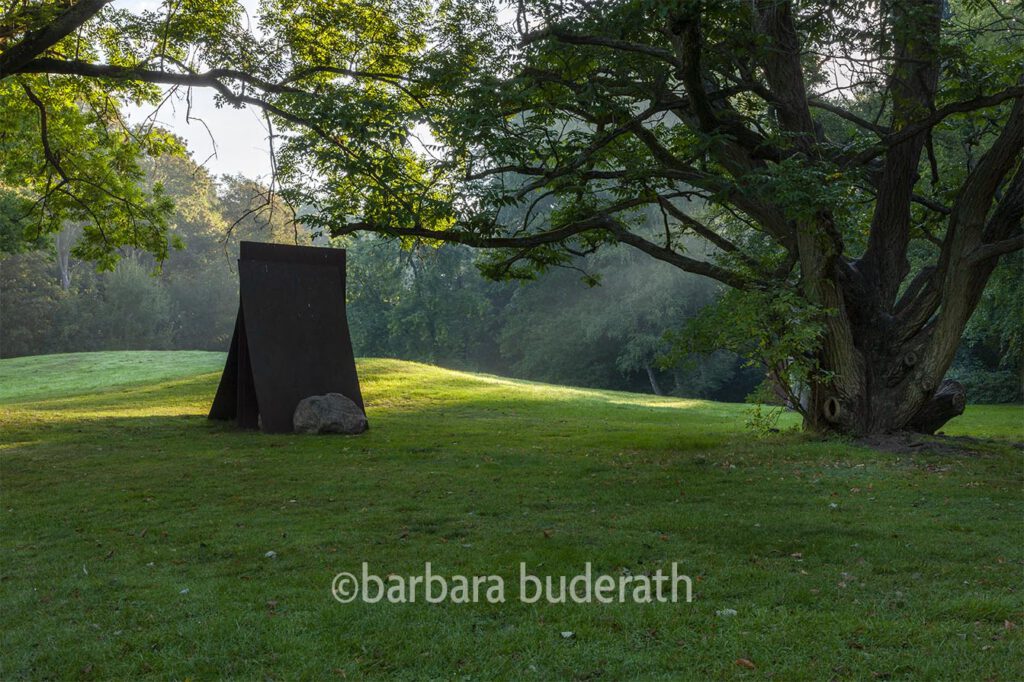 Der Park von Haus Weitmar mit altem Baum im Vordergrund und einem abstrakten Kunstwerk aus rostigen Metallplatten